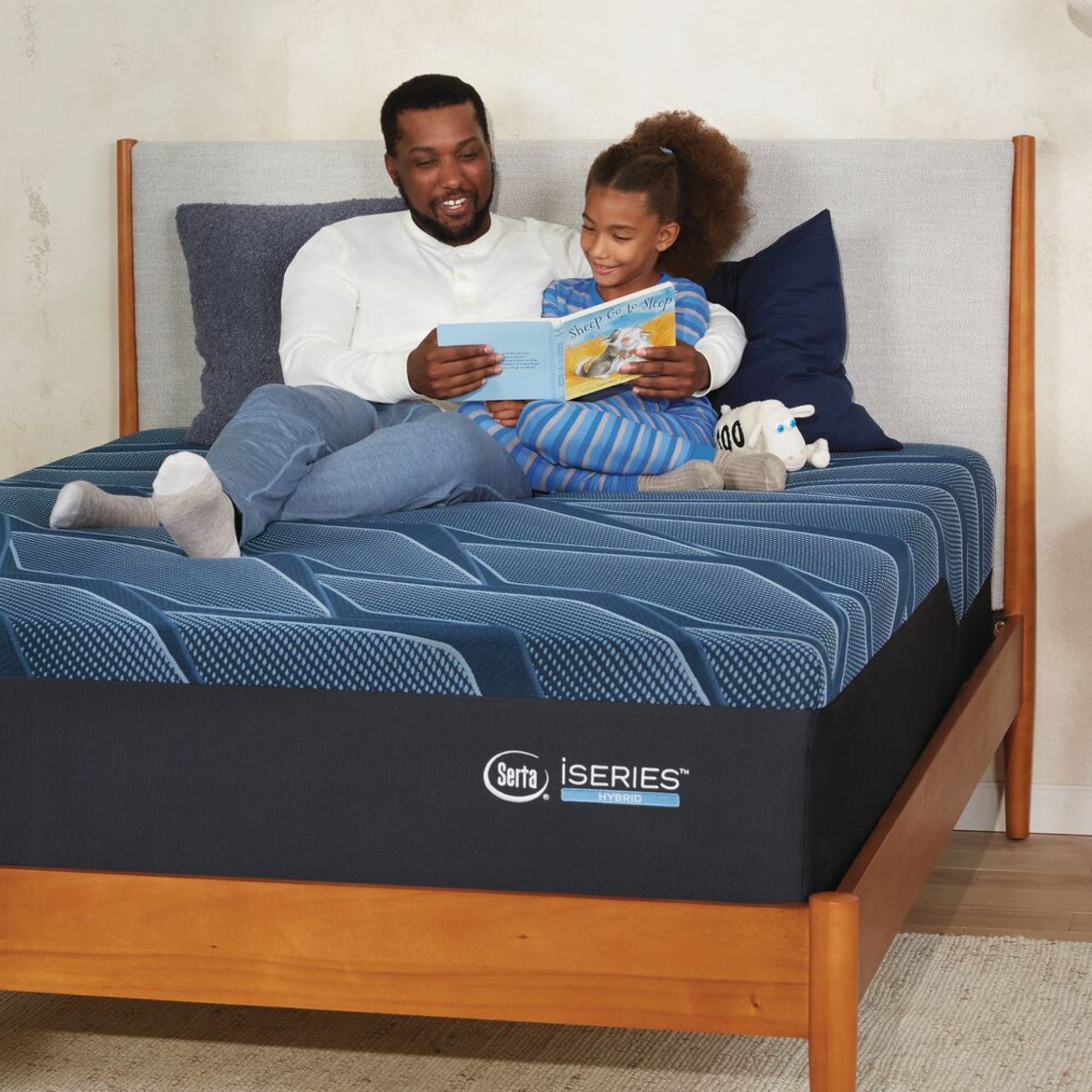 A man and little girl sitting on a Serta iSeries firm blue mattress, placed in a brown bed frame, reading a book while leaning on blue and grey pillows||feel: firm||level: enhanced