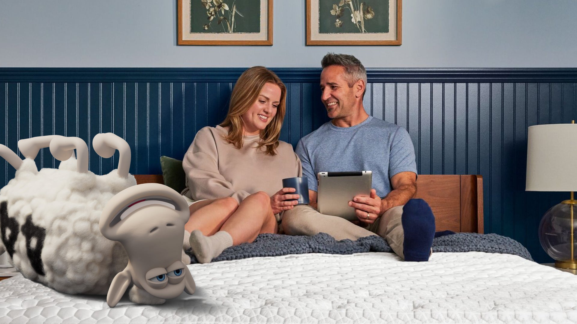 Man and woman sitting on a Serta mattress smiling while looking at an iPad with the number 44 Serta sheep laying on its back at the foot of the bed